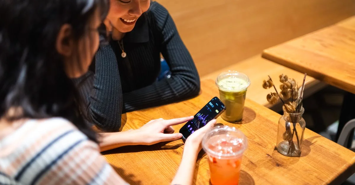 Deux amis prennent un verre dans un café tout en consultant une application boursière sur un smartphone.