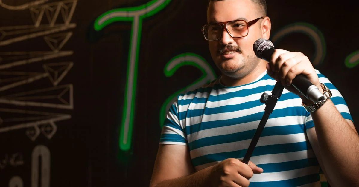 Homme tenant un microphone faisant du stand-up en chemise rayée décontractée sur scène.
