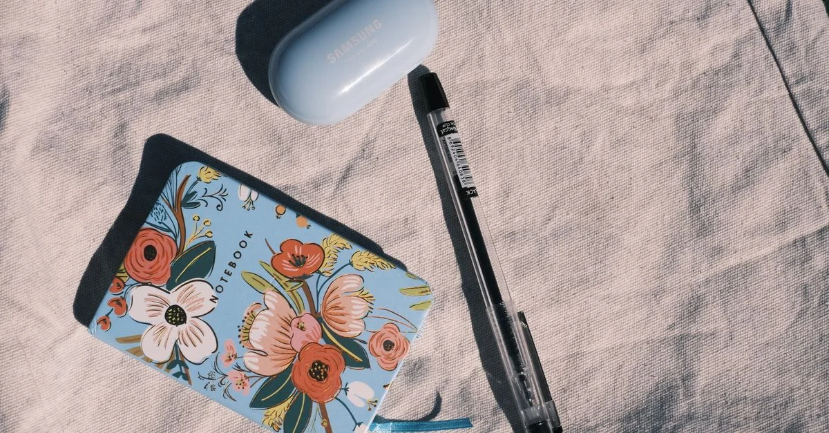 Vue à plat d'un carnet à motifs floraux colorés, d'un stylo et d'une coque Samsung posés sur une surface en tissu, illustrant un style de vie moderne.