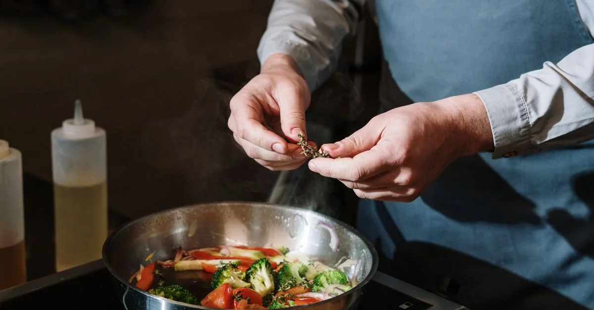 Chef assaisonnant un sauté de légumes dans une poêle avec des herbes fraîches sur une cuisinière.