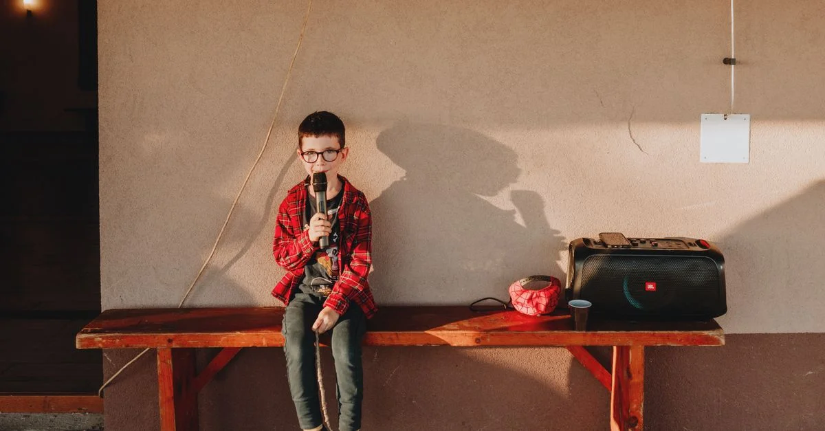 Un jeune garçon chante avec un microphone sur un banc en bois à côté d'un lecteur de musique, profitant d'une journée ensoleillée.